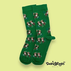 May include: A pair of green socks featuring a repeating pattern of cartoon otters. The otters are depicted in a relaxed pose, floating on their backs. The socks have a ribbed cuff and the brand name "Socks&Rocks" is printed on the lower right.