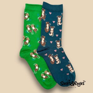 May include: A pair of colorful socks featuring an otter pattern. One sock is green with otters and blue water details, the other is teal with otters and pink hearts. The socks have the text "Socks & Rocks" at the bottom.