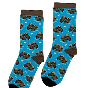 May include: A pair of crew socks in a bright blue colour featuring a repeating pattern of brown sea otters holding hands and small white hearts. The socks have a dark brown cuff at the top and a solid black toe section at the bottom.