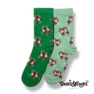 May include: Two pairs of crew socks, one in green and one in light green, featuring a repeating pattern of cartoon otters swimming. The socks have a ribbed cuff and the brand name "Socks&Rocks BERLIN" is printed on the lower right.
