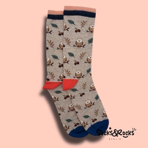Può includere: Un paio di calzini grigi con un motivo ripetuto di gufi appollaiati su rami, foglie e ghiande. I calzini hanno punte rosse, talloni blu e un bordo blu navy e rosa. Il marchio "Socks&Rocks" è stampato in nero.