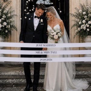 Könnte beinhalten: Ein weißes Zeremonienband mit dem schwarzen Aufdruck JUST MARRIED, MR & MRS TALL und 29.09.2026. Das Band wird vor einem Paar in formeller Hochzeitskleidung gehalten, wobei die linke Person einen schwarzen Smoking und die rechte Person ein weißes, bodenlanges Kleid mit Schleier trägt.