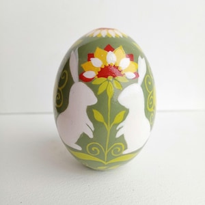Può includere: Un uovo di Pasqua verde con sagome di coniglietti bianchi e un disegno di fiori gialli e rossi.