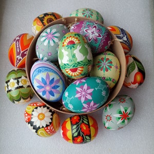 Puede incluir: Una colección de huevos de Pascua pintados a mano en una variedad de colores y diseños, incluyendo patrones florales, formas geométricas y animales. Los huevos están dispuestos en círculo sobre una superficie blanca.
