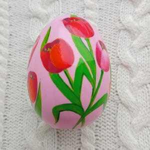 Puede incluir: Un objeto decorativo rosa en forma de huevo con tulipanes rojos pintados a mano con hojas verdes. Los tulipanes tienen centros naranjas. El fondo es una tela blanca texturizada. Este artículo con temática de Pascua es una pieza de decoración del hogar única.