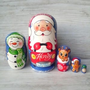 Könnte beinhalten: Ein Set aus fünf handbemalten Matrjoschka-Puppen mit Weihnachtsmotiv. Die Puppen zeigen einen Schneemann, den Weihnachtsmann in einem Auto mit dem Namen "Henry", ein Rentier, einen Lebkuchenmann und eine kleine grüne Puppe. Die Puppen sind in leuchtenden Farben bemalt.