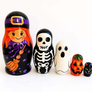 May include: A set of five nesting dolls featuring Halloween characters. The largest is a witch with orange hair and a broom, followed by a skeleton, a white ghost, an orange jack-o'-lantern, and a small black cat with green eyes.