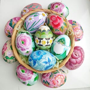 Puede incluir: Una cesta de huevos de Pascua pintados a mano con diseños florales y geométricos. Los huevos son de varios colores, incluyendo rosa, azul, verde y blanco.