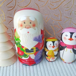 May include: A wooden Christmas tree and three nesting dolls. The largest doll is Santa Claus, and the other two are penguins. The Santa doll is red, white, and black. The penguins are black, white, yellow, and pink.