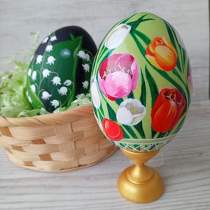 Puede incluir: Dos huevos de Pascua decorativos. Un huevo, en una cesta de mimbre, está pintado con flores blancas de lirios de los valles sobre un fondo verde oscuro. El otro huevo, sobre un soporte dorado, presenta coloridos tulipanes y vegetación sobre un fondo verde claro.
