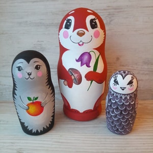 Puede incluir: Tres muñecas rusas de madera pintadas a mano, o muñecas matrioska, que representan una ardilla, un erizo y un búho. La muñeca ardilla es roja con detalles blancos y una flor morada. La muñeca erizo es gris con detalles negros y sostiene una naranja. La muñeca búho es gris con detalles blancos.