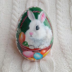 Puede incluir: Huevo de Pascua pintado a mano con un conejito blanco sosteniendo una cesta de huevos coloridos. El conejito está sentado en una cesta verde con un borde rojo. El huevo está decorado con lunares blancos.
