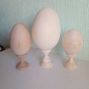 Puede incluir: Tres adornos de madera con forma de huevo de diferentes tamaños. Cada huevo se asienta sobre un pedestal de madera torneada. La madera es de color claro natural con vetas visibles. Los huevos son lisos y simétricos, perfectos para Pascua o exhibiciones decorativas.