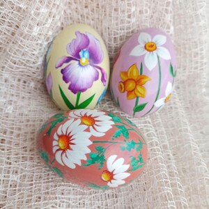 Puede incluir: Tres huevos de Pascua decorados con diseños florales pintados a mano. Un huevo presenta un iris morado, otro una narciso amarillo y una margarita blanca, y el tercero tiene margaritas blancas sobre un fondo rosa.