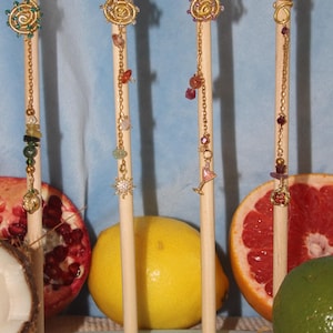 May include: Four gold-toned wire hair coils displayed on wooden sticks. Each coil features intricate wire-wrapped designs with small colourful beads, dangling gold chains, and charms including a sun, a martini glass, and fruit shapes.