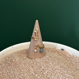 Puede incluir: Un anillo de alambre dorado con un diseño en espiral y una cadena con un pequeño dije transparente de cuatro pétalos y una pequeña cuenta azul claro. El anillo se exhibe sobre un cono de madera, sobre un fondo de arena y un fondo verde oscuro.