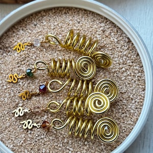 Op de afbeelding: Goudkleurige haarsieraden, waaronder spiraal- en golfontwerpen, met kleine, kleurrijke kralen. De accessoires worden gepresenteerd op een zandbed in een bakje met witte rand. De sieraden zijn ontworpen om als haaraccessoires te worden gebruikt.