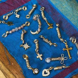 May include: A collection of gold-colored hair accessories, including spiral wraps and charms. Charms include a cross, star, heart, and crescent moon, with some adorned with gemstones. The accessories rest on a blue cloth with red and blue accents.
