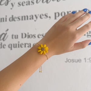 Könnte beinhalten: Ein goldfarbenes Armband mit einem gelben Sonnenblumen-Anhänger mit braunem Zentrum. Das Armband befindet sich an einem Handgelenk mit blauem Nagellack. Der Hintergrund hat spanischen Text, darunter "valiente" und "Josue 1:9."