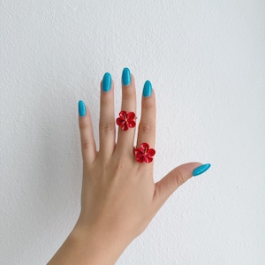 Könnte beinhalten: Zwei rote Blumenringe an einer Hand mit blauem Nagellack.