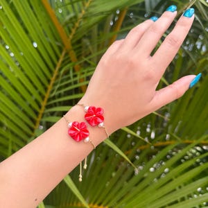 Op de afbeelding: Een goudkleurige armband met twee rode bloemvormige bedels. Elke bloem heeft een witte parel. De armband wordt getoond op een pols, tegen een achtergrond van groene bladeren.