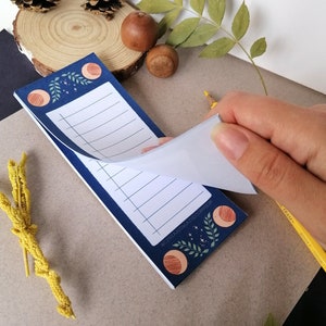Puede incluir: Un bloc de notas azul con un diseño de luna y estrellas. El bloc de notas tiene un interior blanco rayado y está doblado por la mitad. El bloc de notas está sobre una superficie de madera con una bellota marrón y una piña.