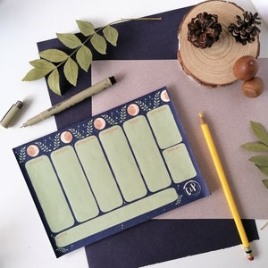 Pode incluir: Um bloco de notas de planejador semanal com uma borda azul escura com um design celestial com luas e folhas. O bloco de notas tem seções verdes para cada dia da semana, uma seção de notas e um lápis amarelo.