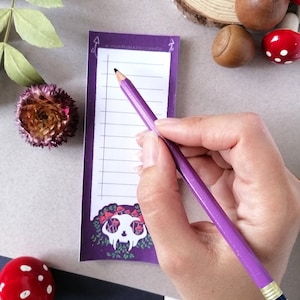 May include: Purple notepad with a skull and mushrooms illustration. The notepad has lined paper and is ideal for writing down lists or notes. A purple pencil is being used to write on the notepad.