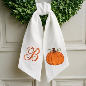 May include: A white fabric hanging decoration featuring an embroidered orange pumpkin on the right side and a large, cursive orange letter B on the left side. The fabric is knotted at the top and hangs against a green leafy wreath background.