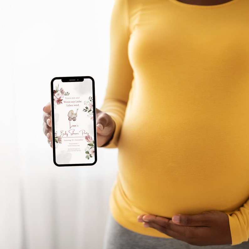 May include: A person holding a smartphone displaying a baby shower invitation with a floral design and the text "Wenn aus Liebe Leben wird..." and "Lena's Baby Shower Party".