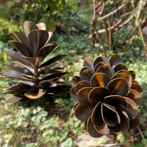 May include: Two metal plant sculptures with a rustic finish. One is a layered succulent, the other a pinecone shape. The sculptures are brown with orange highlights, set against a blurred green background of foliage.
