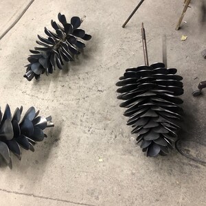 May include: Three metal pine cone sculptures, each with a different size and shape. The sculptures are made of individual metal pieces that are welded together to form the shape of a pine cone. The sculptures are all black in colour and have a rustic, industrial look.