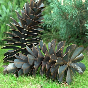 May include: Two large, dark brown metal pine cones rest on green grass, with evergreen trees in the background. The metal pine cones have detailed, overlapping scales and a weathered appearance. The sculpture is a decorative garden accent.