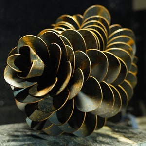 May include: A metal sculpture resembling a pine cone, made of overlapping, rusty, circular pieces. The sculpture is displayed on a dark, textured surface.