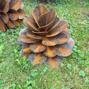 May include: A rusty metal sculpture of a pine cone with overlapping petals. The sculpture is brown and orange with a weathered look. It is sitting on a bed of green grass.
