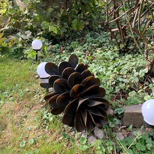 May include: A large, dark brown metal garden sculpture, shaped like a flower or pinecone, sits amidst green grass and foliage. Two white globe lights are visible, adding to the decorative outdoor setting.