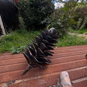 Puede incluir: Una escultura decorativa con forma de piña grande, construida con múltiples placas metálicas oscuras superpuestas dispuestas en espiral. El objeto presenta un tallo central y un acabado metálico oscuro y liso en toda su estructura.
