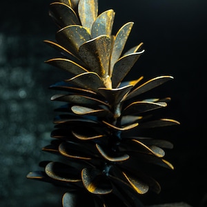 May include: A close-up of a metal sculpture resembling a pinecone. The sculpture has layered, petal-like shapes in dark grey and brown, with golden accents. The background is a dark, blurred texture. The sculpture is likely made of iron.