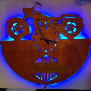 May include: Wooden motorcycle clock with blue LED backlighting. The clock features a motorcycle silhouette above a clock face, with the words "MOTOR CYCLES" cut out at the bottom. The clock hands are black.