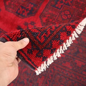 Puede incluir: Una alfombra roja y azul marino con un dise&ntilde;o geom&eacute;trico detallado. La alfombra est&aacute; parcialmente doblada, revelando la parte inferior y el fleco blanco. Los colores intensos y los patrones intrincados sugieren un estilo tradicional u oriental.