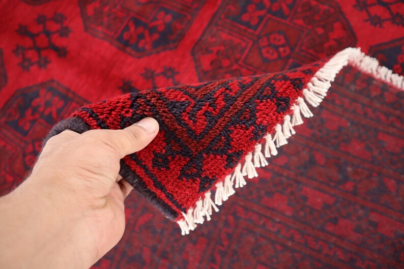 Puede incluir: Una alfombra roja y azul marino con un dise&ntilde;o geom&eacute;trico detallado. La alfombra est&aacute; parcialmente doblada, revelando la parte inferior y el fleco blanco. Los colores intensos y los patrones intrincados sugieren un estilo tradicional u oriental.