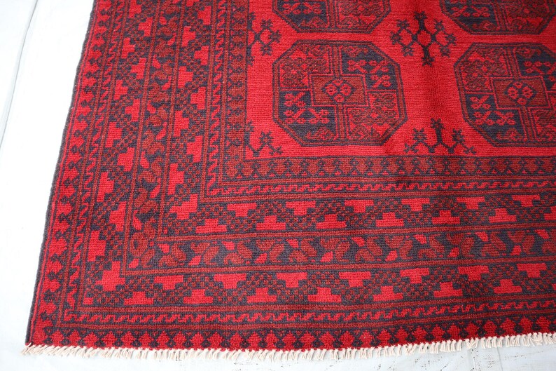 Puede incluir: Secci&oacute;n de una alfombra estampada en rojo y negro. La alfombra presenta dise&ntilde;os geom&eacute;tricos y un borde con un patr&oacute;n repetitivo. La combinaci&oacute;n de colores de la alfombra es principalmente roja con acentos negros y tiene un borde con flecos.