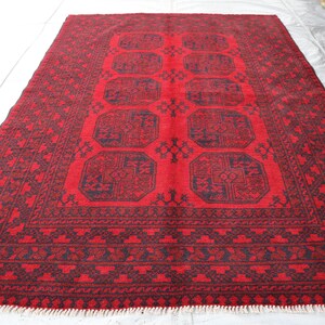 Puede incluir: Una alfombra rectangular roja con un dise&ntilde;o geom&eacute;trico. La alfombra presenta un campo central rojo con formas octogonales azul oscuro repetidas. Un borde azul oscuro enmarca la alfombra, y la alfombra tiene flecos en los extremos cortos.