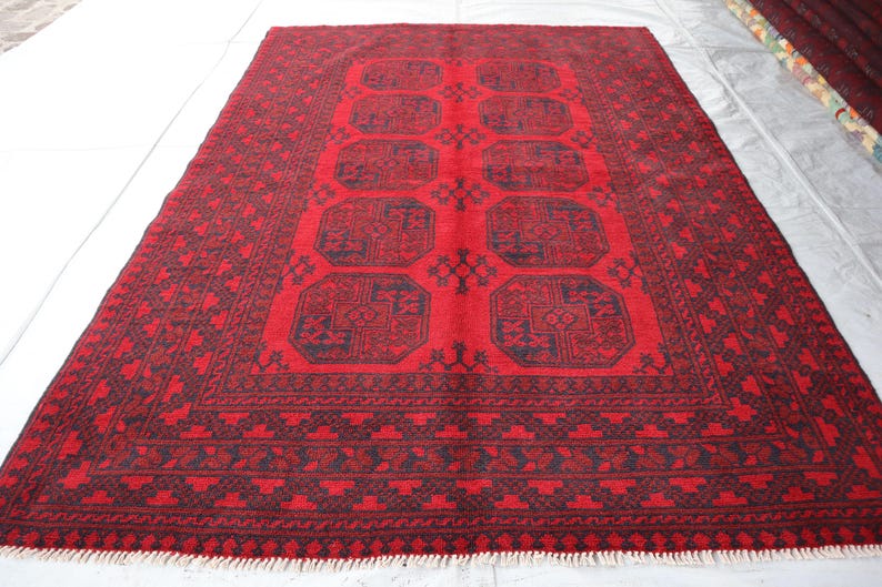 Puede incluir: Una alfombra rectangular roja con un dise&ntilde;o geom&eacute;trico. La alfombra presenta un campo central rojo con formas octogonales azul oscuro repetidas. Un borde azul oscuro enmarca la alfombra, y la alfombra tiene flecos en los extremos cortos.