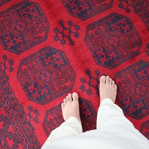 Puede incluir: Una vibrante alfombra roja con intrincados patrones geom&eacute;tricos en azul marino y rojo oscuro. La alfombra presenta un dise&ntilde;o central de formas octogonales repetidas y motivos florales. La imagen muestra los pies de una persona y pantalones blancos sobre la alfombra.