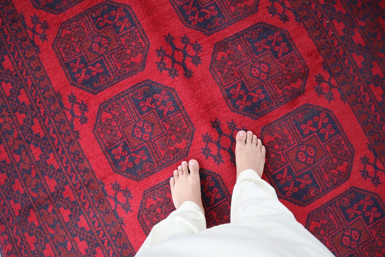 Puede incluir: Una vibrante alfombra roja con intrincados patrones geom&eacute;tricos en azul marino y rojo oscuro. La alfombra presenta un dise&ntilde;o central de formas octogonales repetidas y motivos florales. La imagen muestra los pies de una persona y pantalones blancos sobre la alfombra.