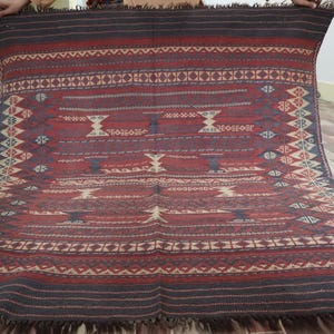 May include: A rectangular, hand-woven rug with a geometric pattern in shades of red, burgundy, and cream. The rug features a central design with repeating motifs and a dark border with fringe along the edges. The rug is displayed against a light-colored wall.