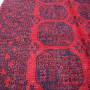 Puede incluir: Una alfombra roja y azul marino estampada. La alfombra presenta un dise&ntilde;o geom&eacute;trico repetitivo con formas octogonales y detalles intrincados. El borde tiene un patr&oacute;n repetitivo de formas azul marino. La alfombra est&aacute; hecha de un material tejido.