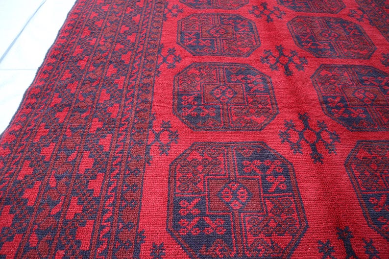 Puede incluir: Una alfombra roja y azul marino estampada. La alfombra presenta un dise&ntilde;o geom&eacute;trico repetitivo con formas octogonales y detalles intrincados. El borde tiene un patr&oacute;n repetitivo de formas azul marino. La alfombra est&aacute; hecha de un material tejido.