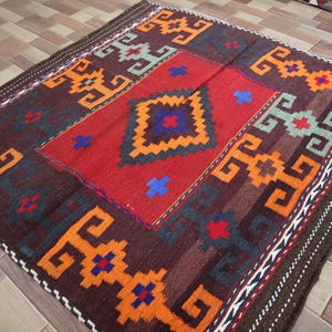 May include: A traditional Turkish rug with a geometric pattern in shades of red, orange, blue, and brown. The rug has a fringe border and is woven with wool.